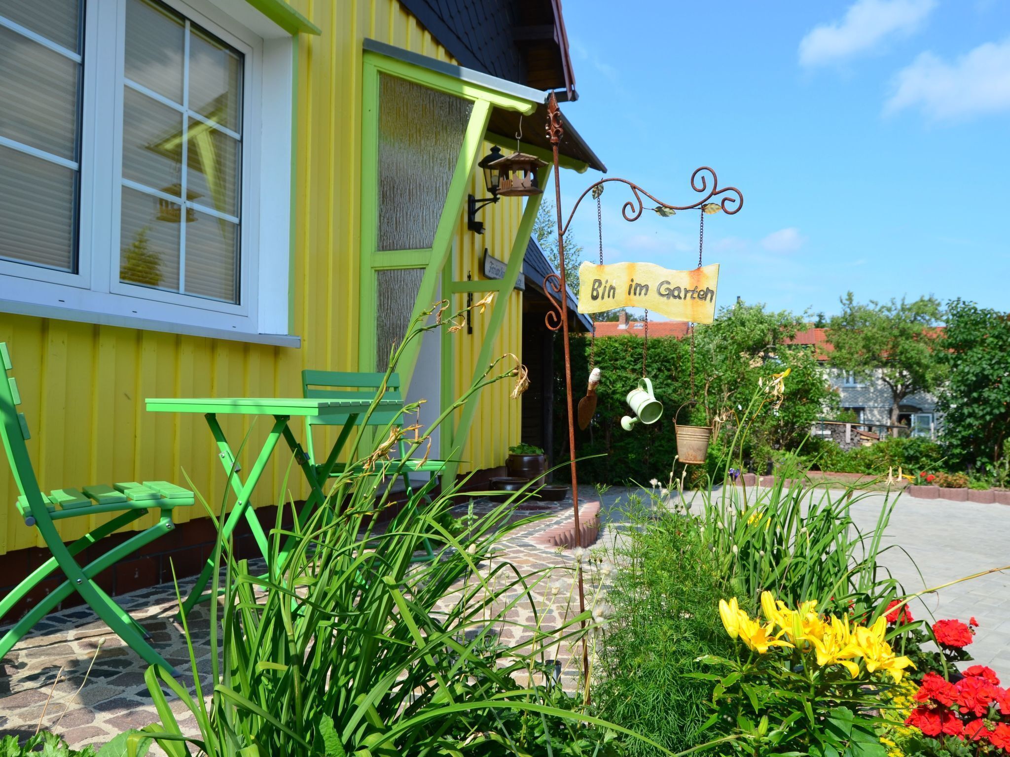 A yellow building with a green door and windows, a sign reading "Bin im Garten", and a garden with plants and flowers.