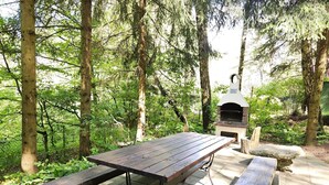 An outdoor wooden picnic table with benches, surrounded by a forested area.
