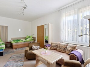 Uma sala de estar acolhedora com um sofá, uma mesa de centro de madeira e uma poltrona. Há um espelho na parede e uma janela com cortinas leves.