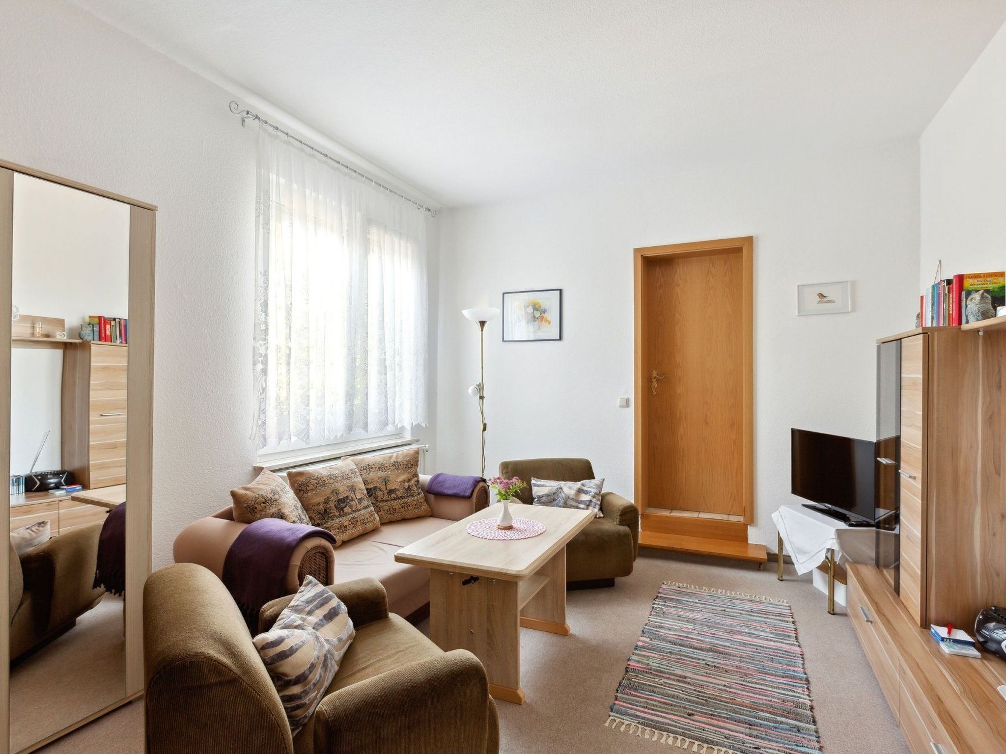 Un salon moderne comprenant un canapé, un fauteuil, une table basse et un téléviseur. On y trouve également une porte en bois, un miroir et une bibliothèque remplie de livres.
