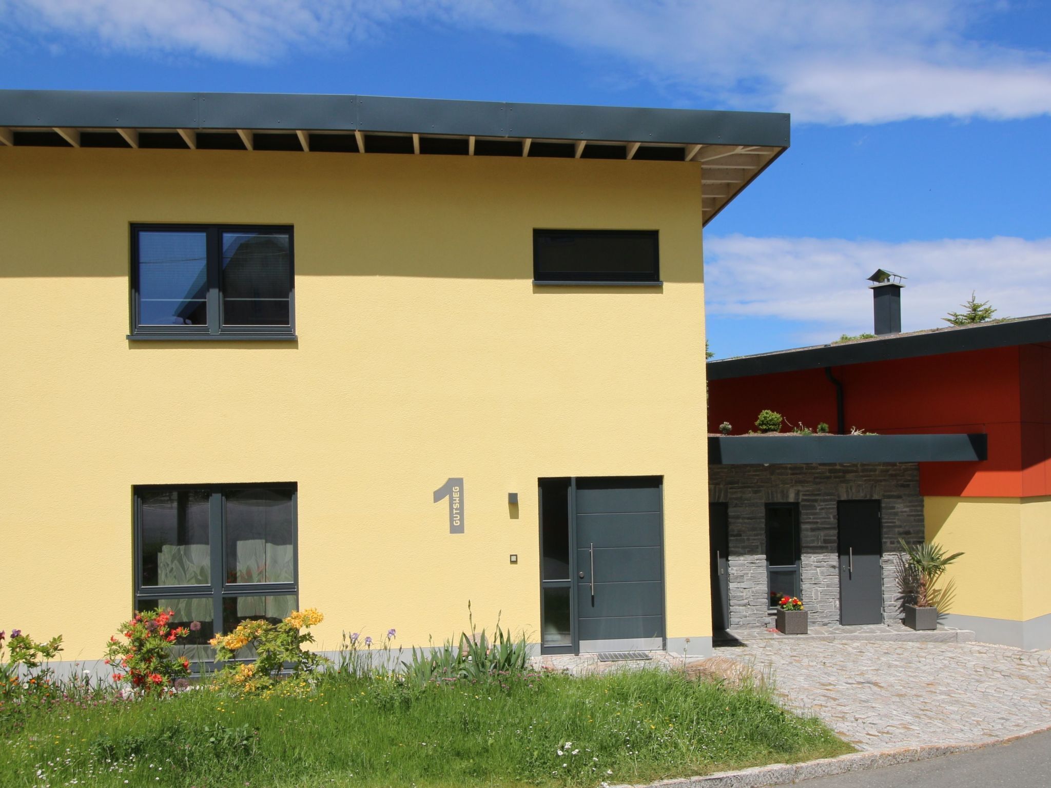 A modern two-story building with a yellow facade, a stone entrance, and a small garden area with flowers.