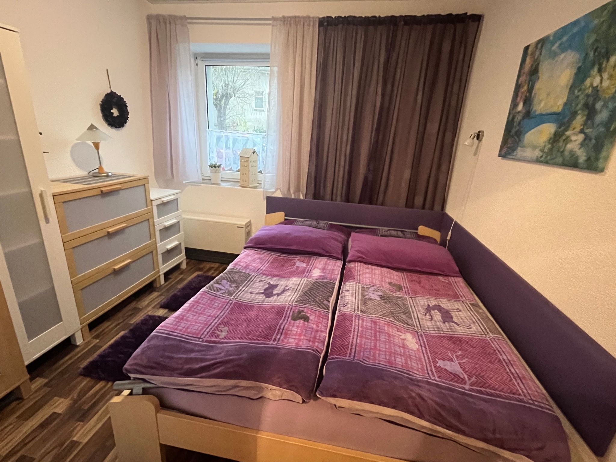 Ein Schlafzimmer mit Bett, Schminktisch, einem Fenster mit Vorhängen und einem Bild an der Wand.