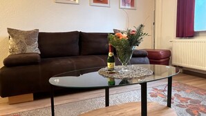 Un salón con un sofá de cuero marrón, una mesa de centro de cristal con una botella y flores, y un radiador.