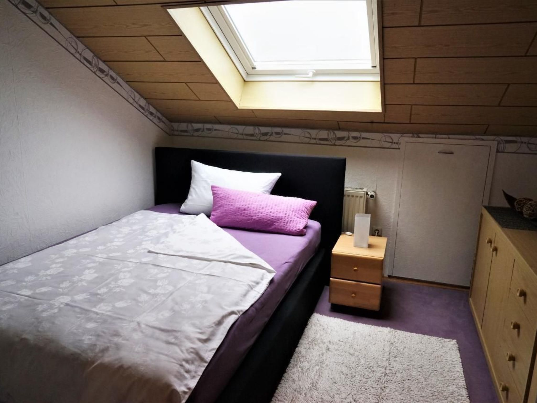 A bedroom with a skylight, a bed with a white and purple bedspread, a nightstand with a lamp, and a dresser.