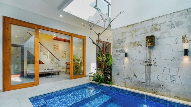A modern indoor pool with clear blue tiles, a wooden staircase, and a glass door leading to a seating area with a sofa and a table. - 3-bedroom Private pool villa City Sanur (Denpasar)