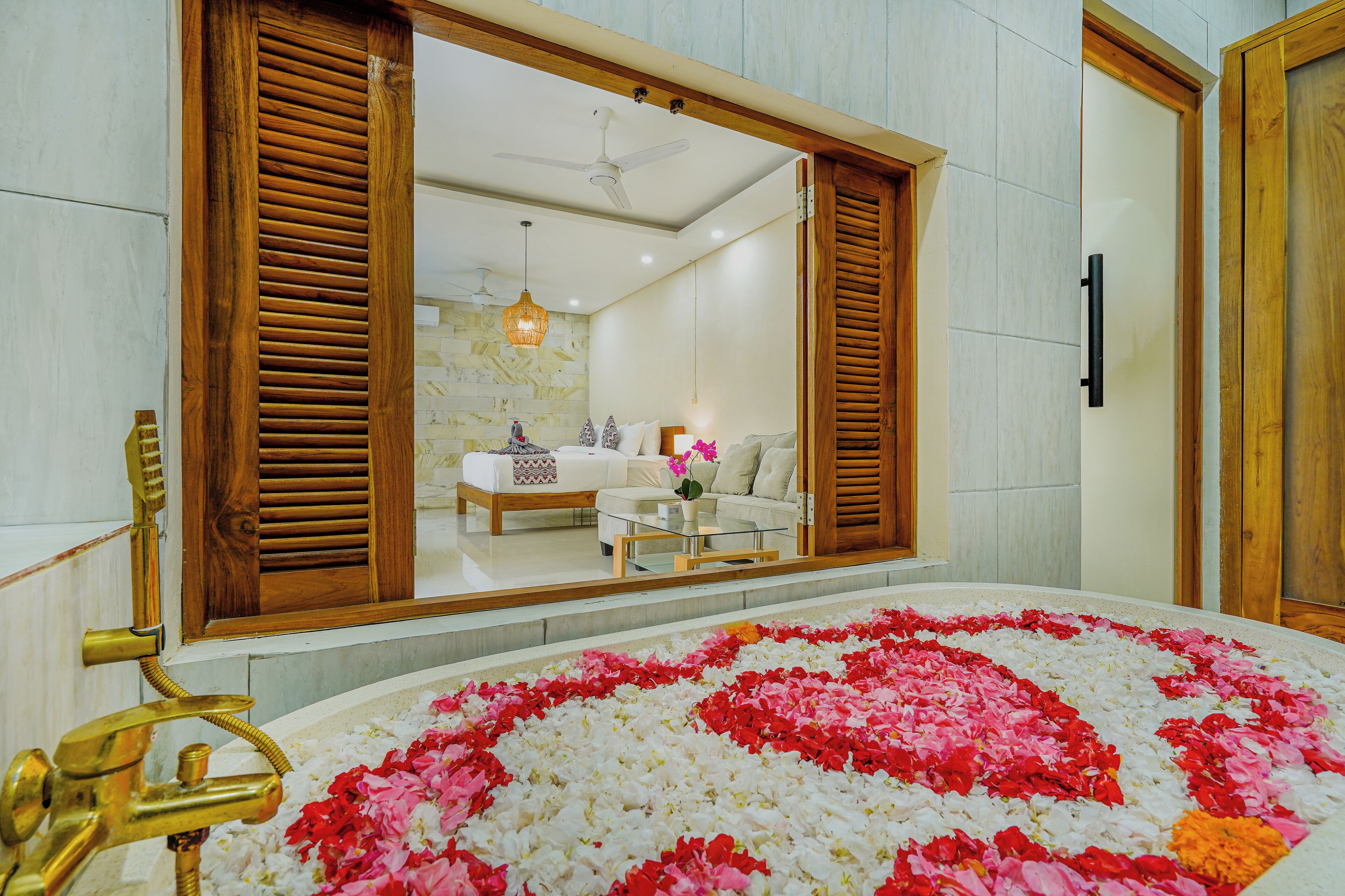 Un baño con un espejo grande, contraventanas de madera y una bañera rodeada de pétalos de rosa.