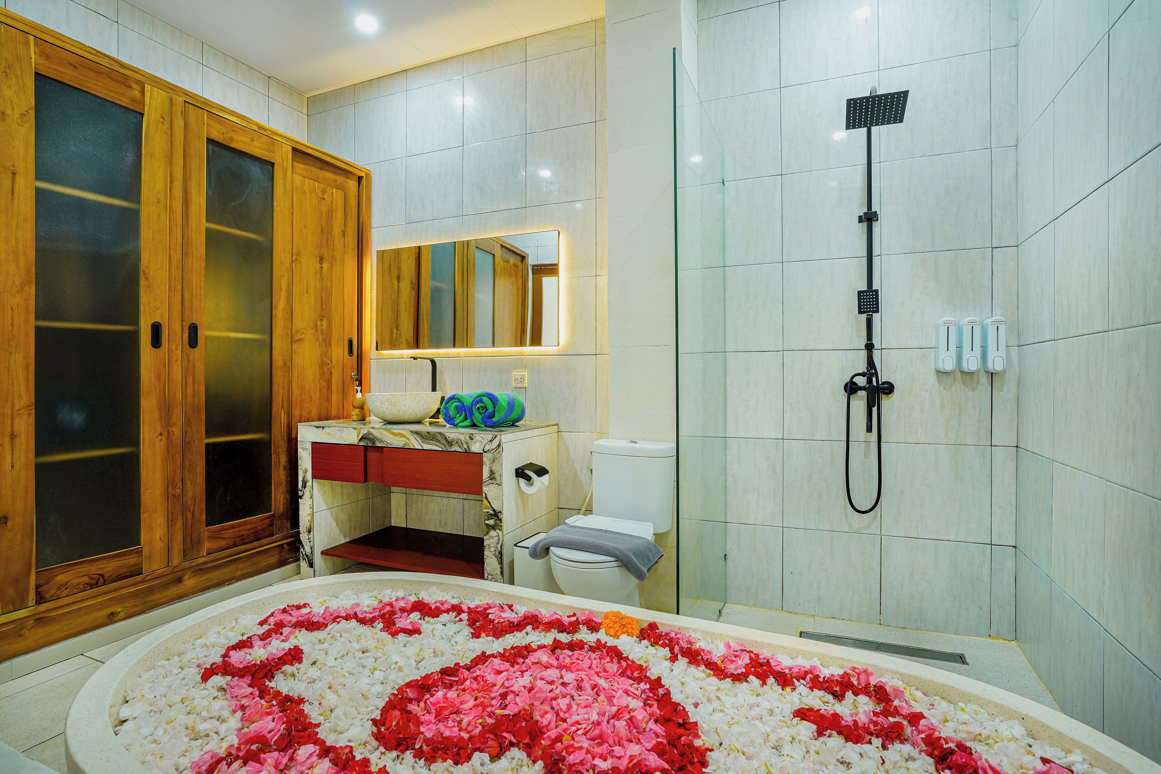 Un baño con un espejo grande, armarios de madera, una ducha y una bañera con pétalos de rosa.