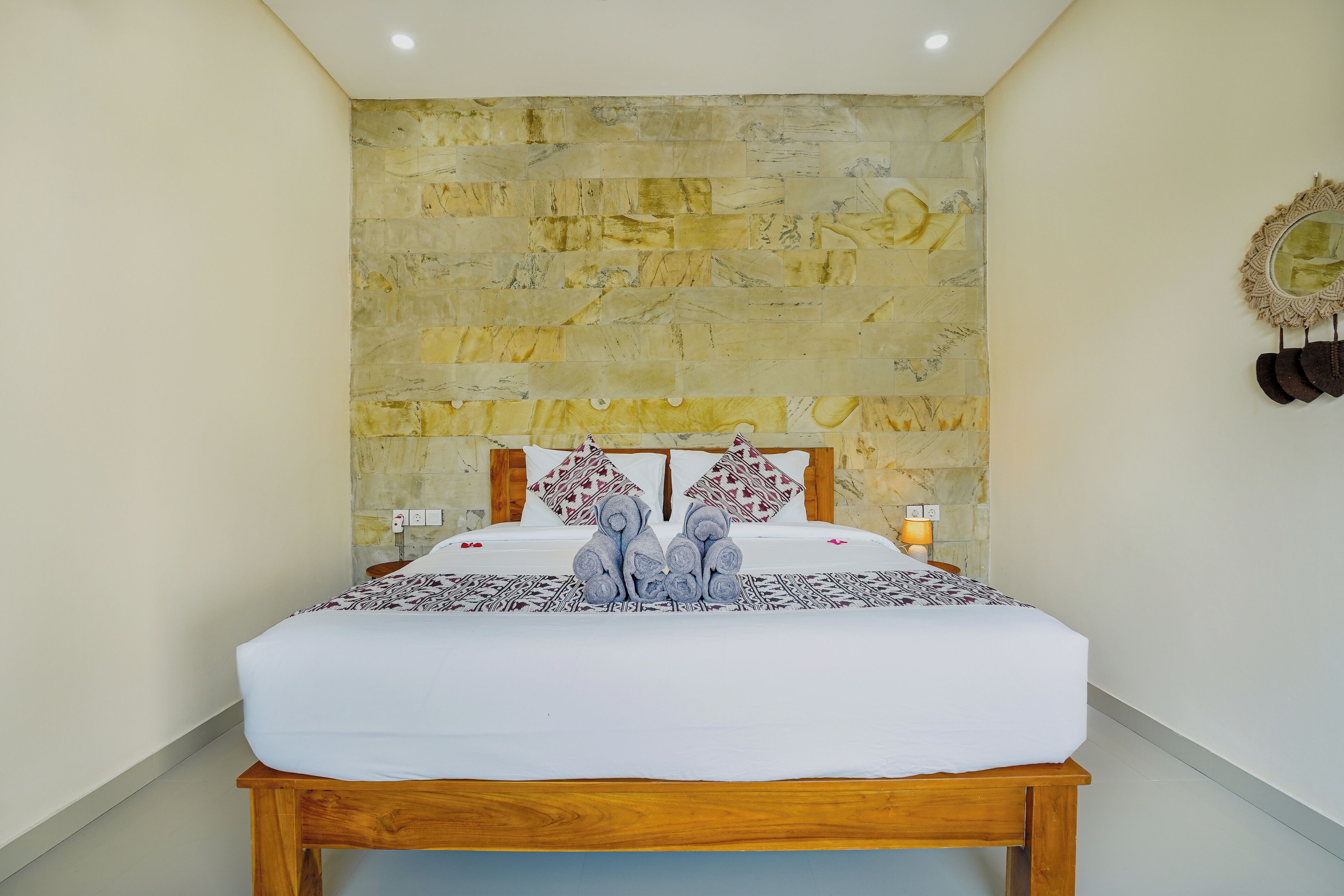 Una cama bien tendida con colcha y almohadas estampadas, cabecera de madera y pared de piedra al fondo.