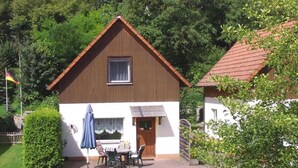 A two-story house with a brown roof and white walls, a small window, and a door. There is a patio with a table and chairs, and two striped lounge chairs. - Haus im Grünen by Interhome (Marktgraitz)