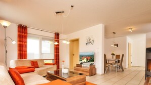 Ein modernes Wohnzimmer mit Sofa, Couchtisch und Fernseher. Es gibt einen Essbereich mit Stühlen und Tisch. Der Raum verfügt über einen Kamin und ein großes Fenster.