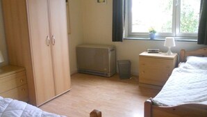 Ein Schlafzimmer mit Holzschrank, Nachttisch mit Lampe, Heizung und Fenster mit Blick ins Grüne.