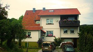 Ein zweistöckiges Haus mit roten Ziegeldach, einem Balkon mit Blumen und zwei geparkten Autos davor.