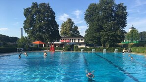 Une piscine à l’eau d’un bleu cristallin, entourée d’arbres et bordée d’un bâtiment à la façade blanche.