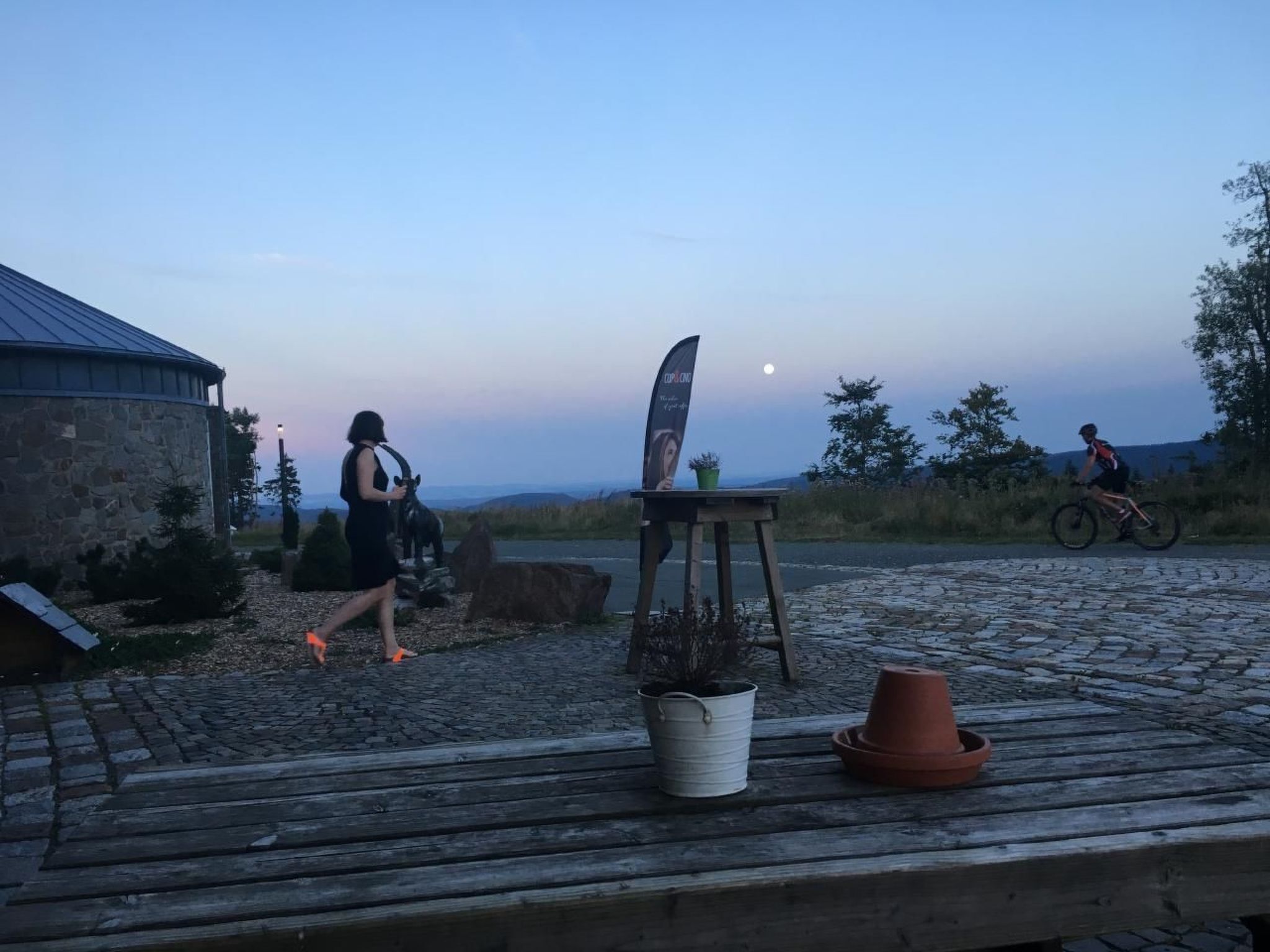 Eine rustikale Outdoor-Szene mit einem Holztisch, einem Steinhaus und einem Radfahrer im Hintergrund.
