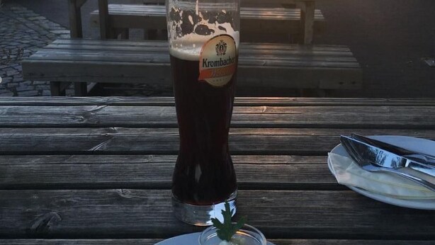 Une table en bois avec un verre de bière brune, une assiette de nourriture et un banc à l’arrière-plan.