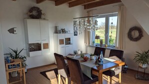 Een eethoek met een houten tafel en stoelen, een wit kastje met klokken, en uitzicht op het landschap door grote ramen.