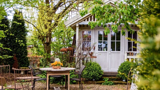 Una casa de madera rústica con un porche, rodeada por un jardín que incluye una mesa y sillas, y un pequeño estanque con una estatua.