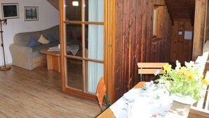 Interior acolhedor em madeira com uma porta de vidro deslizante que dá para uma sala de estar, uma mesa de jantar posta com um arranjo de flores e um sofá com almofadas.