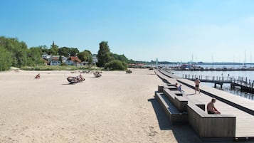 Ein sandiger Strand mit einer Holzstege, der sich in ruhiges Wasser erstreckt, umgeben von Grünflächen und Häusern im Hintergrund.