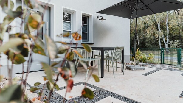 Een terras met een tafel, stoelen, een grote parasol en uitzicht op een wit gebouw met ramen.
