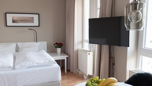Une chambre d’hôtel avec un lit, une table de chevet ornée d’un vase de fleurs, un radiateur, un téléviseur fixé au mur et une table sur laquelle se trouve un panier de fruits.