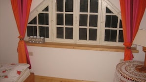 Ein Schlafzimmer mit einem hölzernen Bett, einem Fenster mit weißen Lamellen und einem kleinen runden Tisch mit einer gehäkelten Tischdecke.