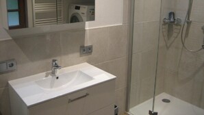 Ein Badezimmer mit weißem Waschbecken, einer Duschkabine aus Glas und einem Spiegel, der einen Bereich für die Wäsche zeigt.
