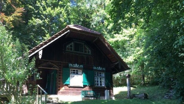 Eine rustikale Holzhütte mit einem steilen Satteldach, umgeben von üppigem Grün.