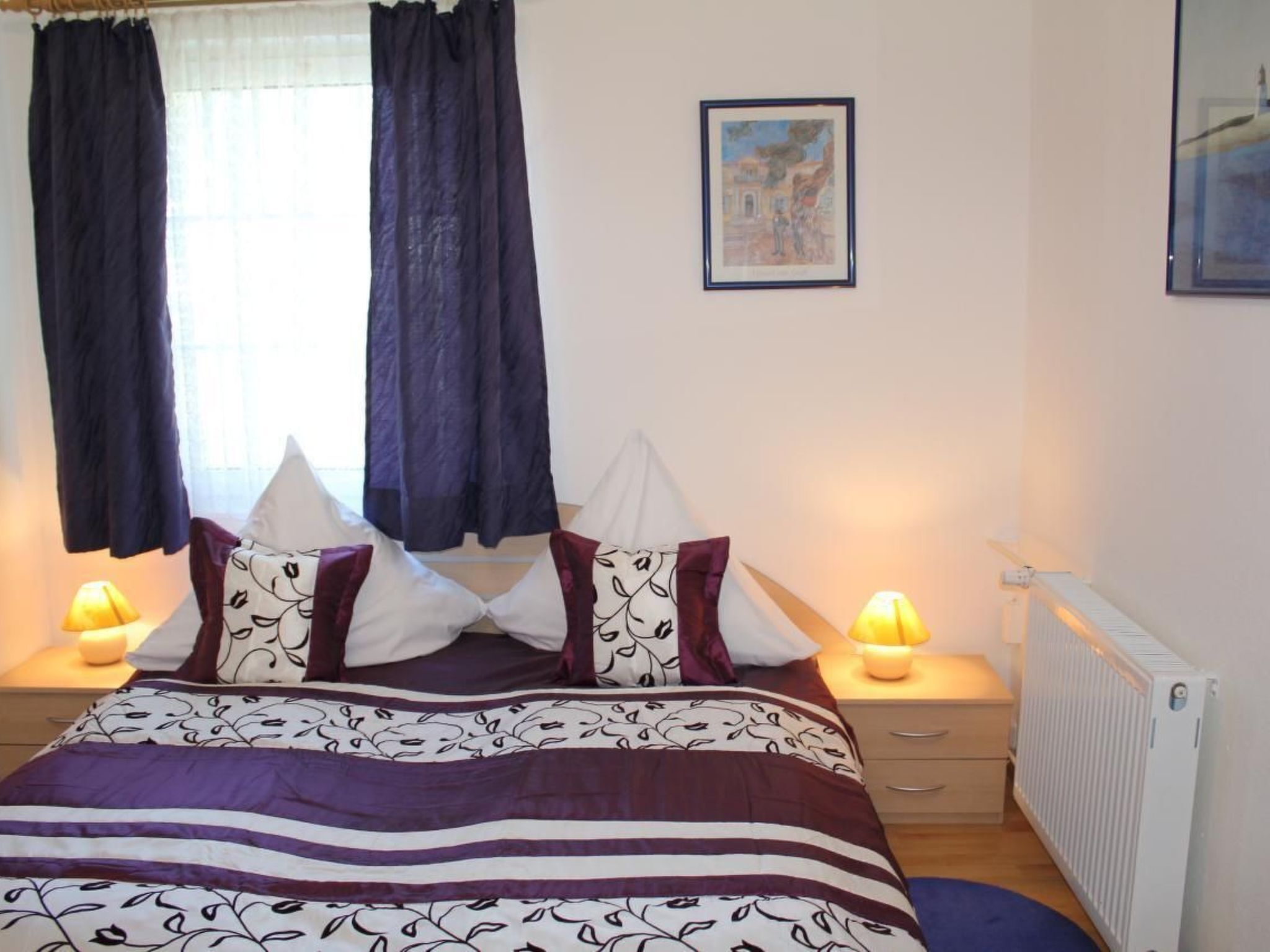 A bedroom with a bed, two bedside lamps, a radiator, and a framed picture on the wall.
