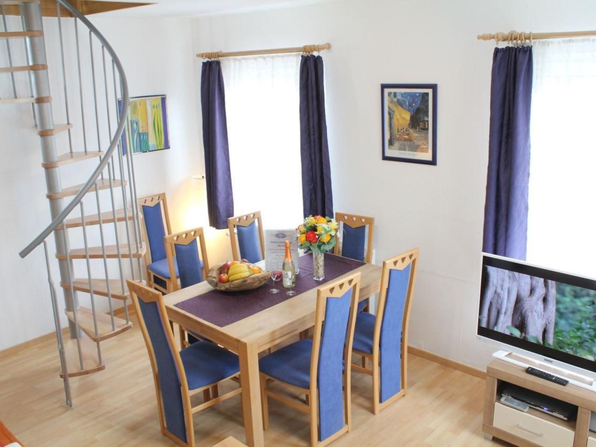 Un comedor con una mesa de madera, sillas azules, un frutero, una botella y un televisor sobre un mueble.