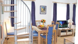 A modern living room with a spiral staircase, a dining area with blue chairs, a TV, and a wooden table with a fruit bowl. - Schöne Ferienwohnung in Daun mit Garten by Interhome (Daun)