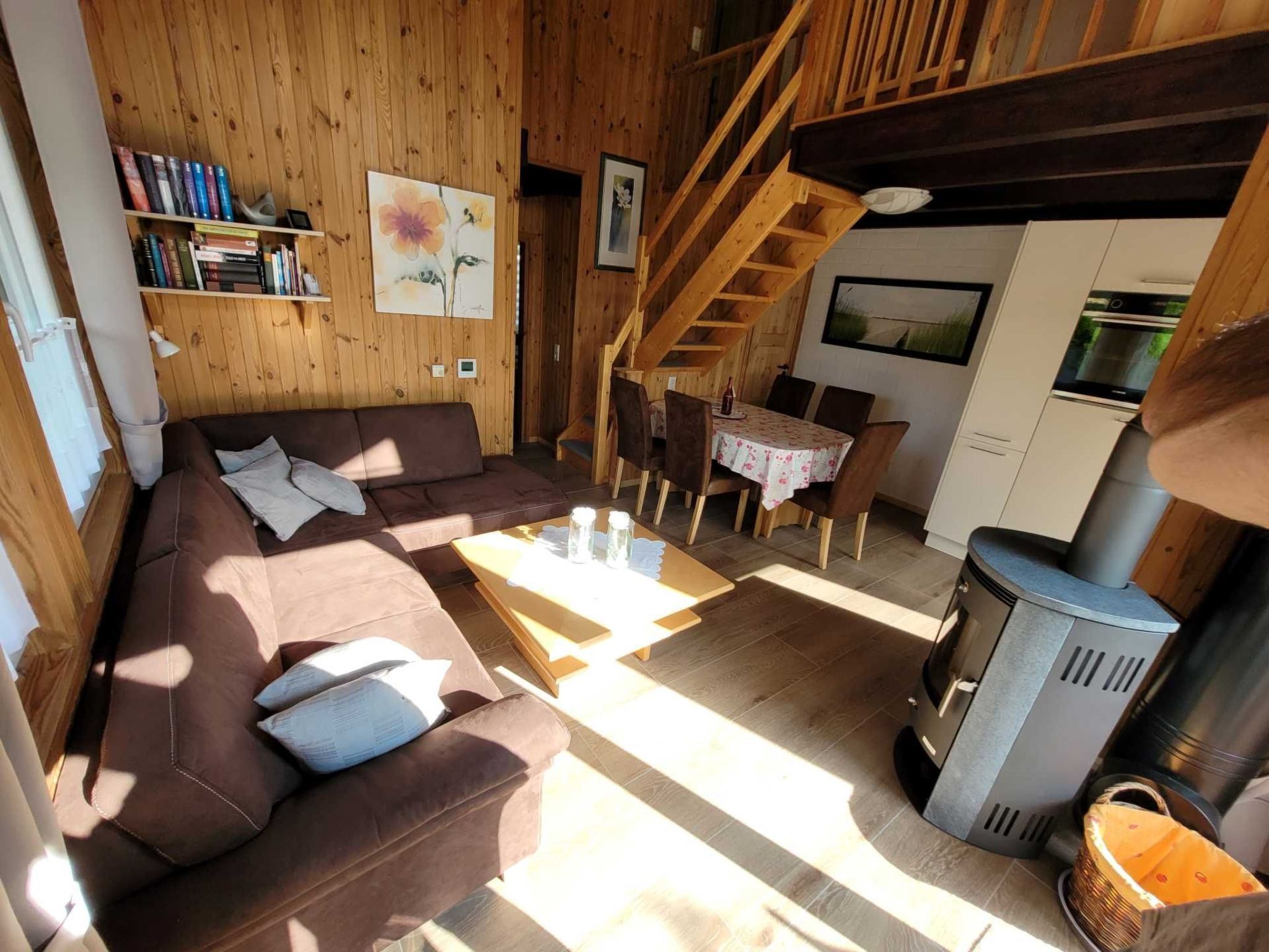 A cozy cabin interior with a wooden interior, a sofa, a dining table, chairs, a staircase, and a wood-burning stove.