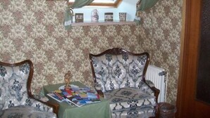 Ein Zimmer mit gemustertem Wandpapier, einem grünen Tisch mit Zeitschriften, zwei Polsterstühlen und einer Holzkassette mit Dekorationen.