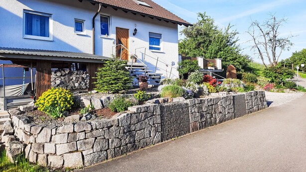 Une maison blanche avec un toit en tuiles, un mur en pierre et un jardin où poussent différentes plantes et fleurs.