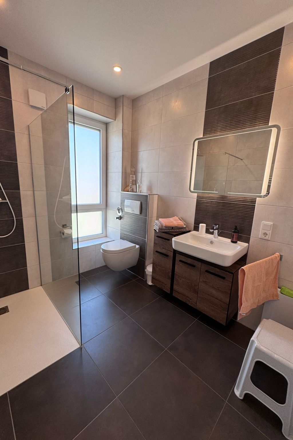 Een moderne badkamer met een douchecabine van glas, een toilet, een wastafel met spiegel en een klein krukje.