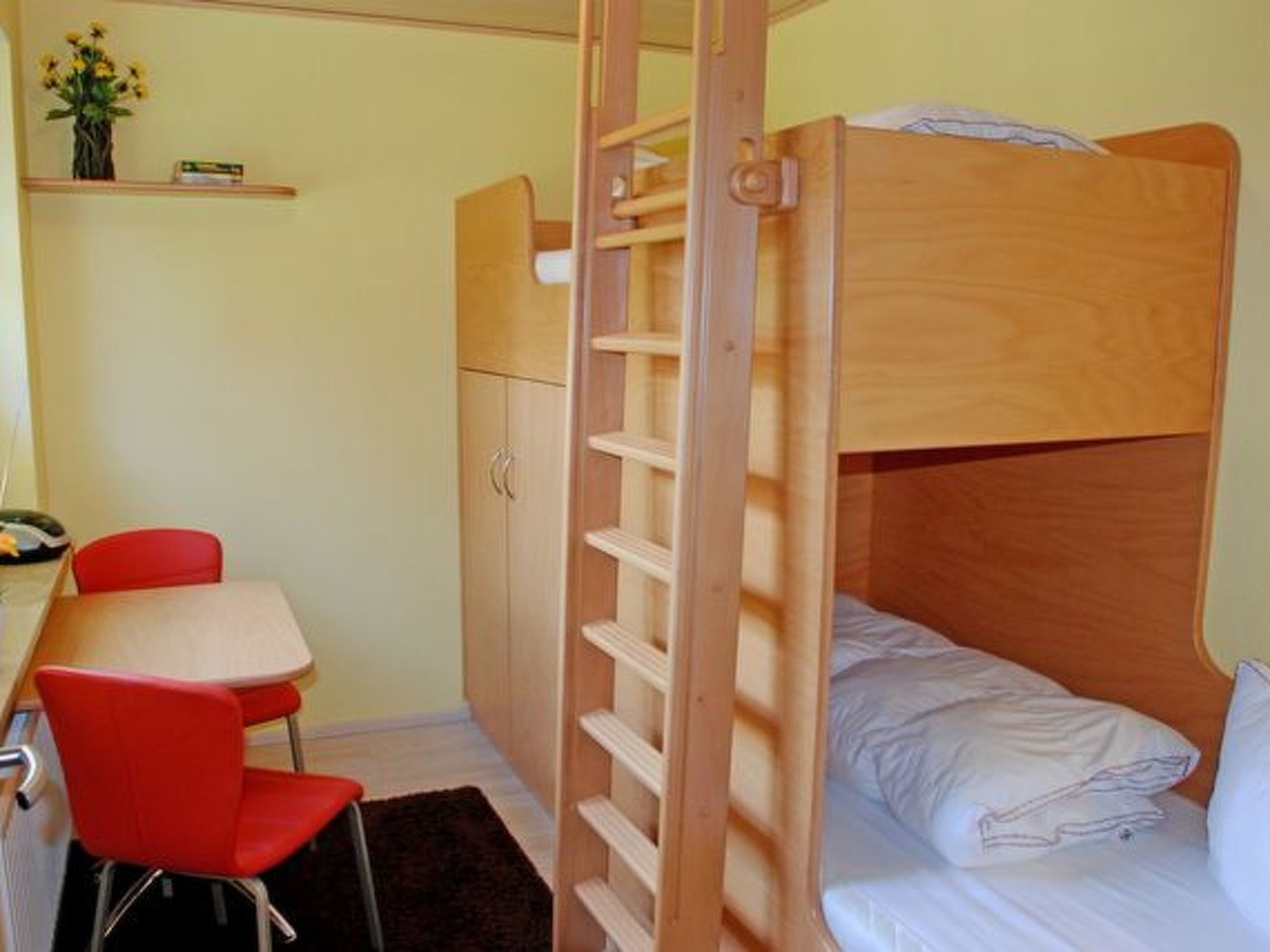 Ein kleines Zimmer mit Etagenbett, Schreibtisch mit Stuhl und einer kleinen Ablage mit Vase.