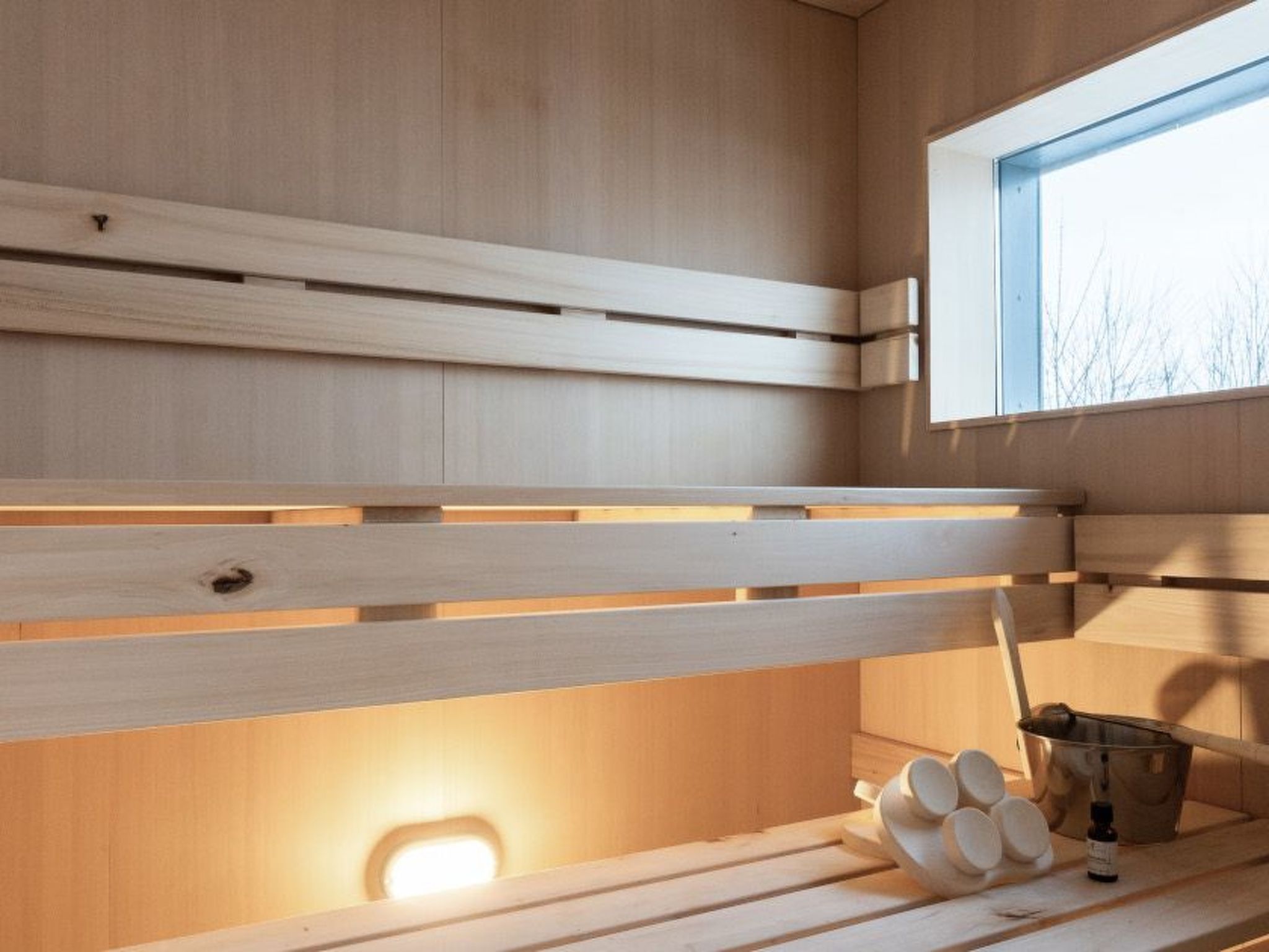 A sauna with wooden benches and a window, a bucket, and a bottle.