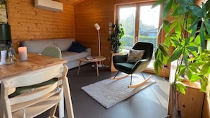 A cozy cabin interior with a wooden ceiling, a sofa, a dining table with chairs, a rocking chair, and a large window with a view of greenery. - in Atzenhain mit Großer Terrasse by Interhome (Atzenhain)