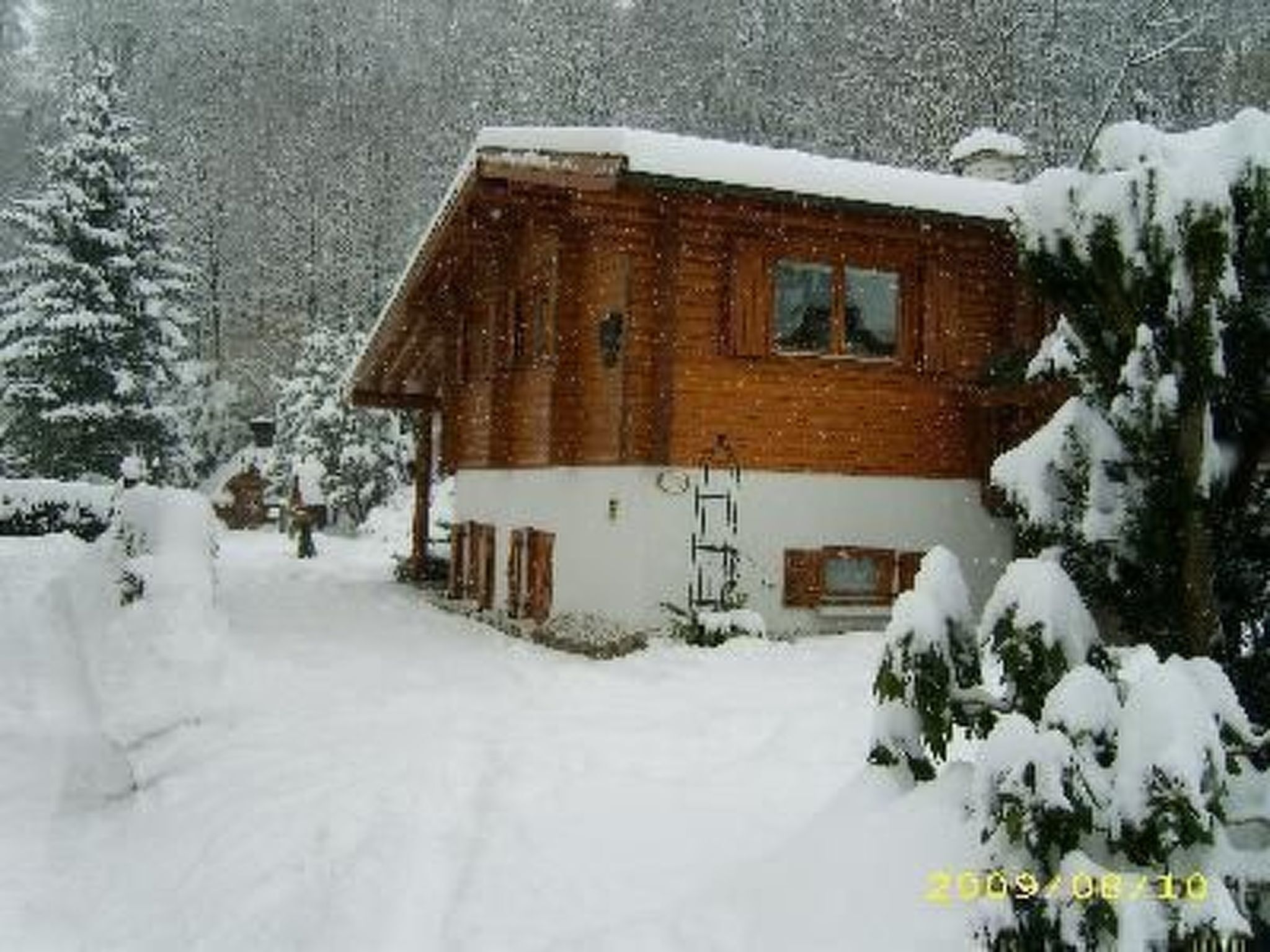 Une cabane en bois, dont le toit est recouvert de neige et dont la cheminée est bien visible, entourée d’arbres chargés de neige.