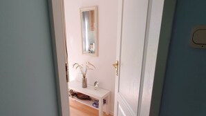 Een gang met een witte deur, een klein wit bankje en een spiegel aan de muur.