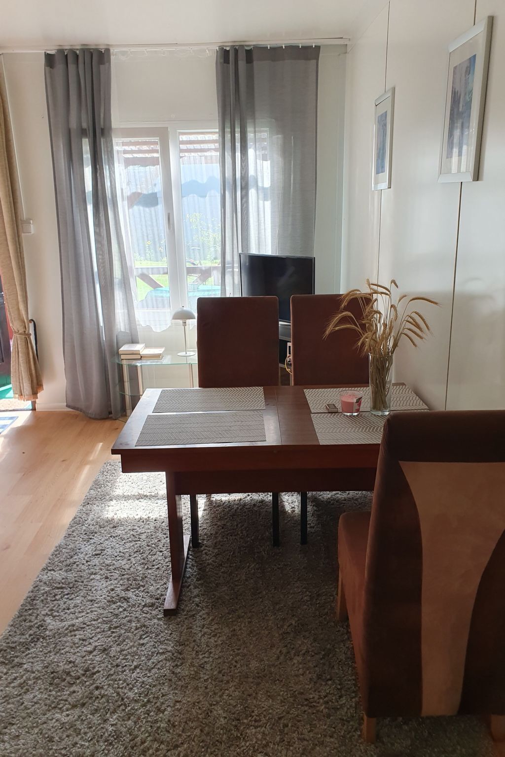 A living room with a wooden dining table, two chairs, a TV, and a view of a garden through large windows.