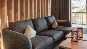Uma sala de estar moderna com um sofá cinzento, duas almofadas, uma mesa de centro de madeira e uma janela grande com cortinas.