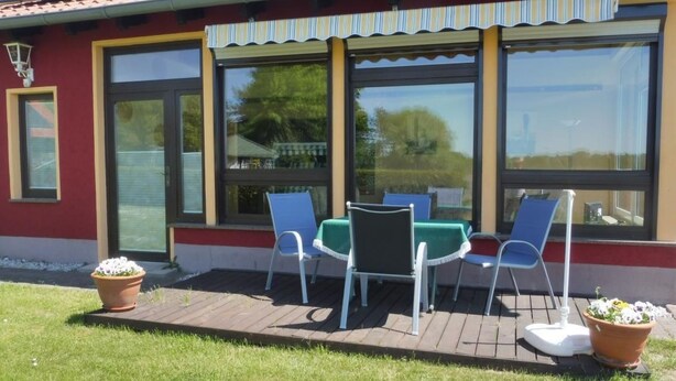 Rakennuksessa on suuret ikkunat, terassi, jossa on tuoleja, ja ruukuissa olevia kasveja.