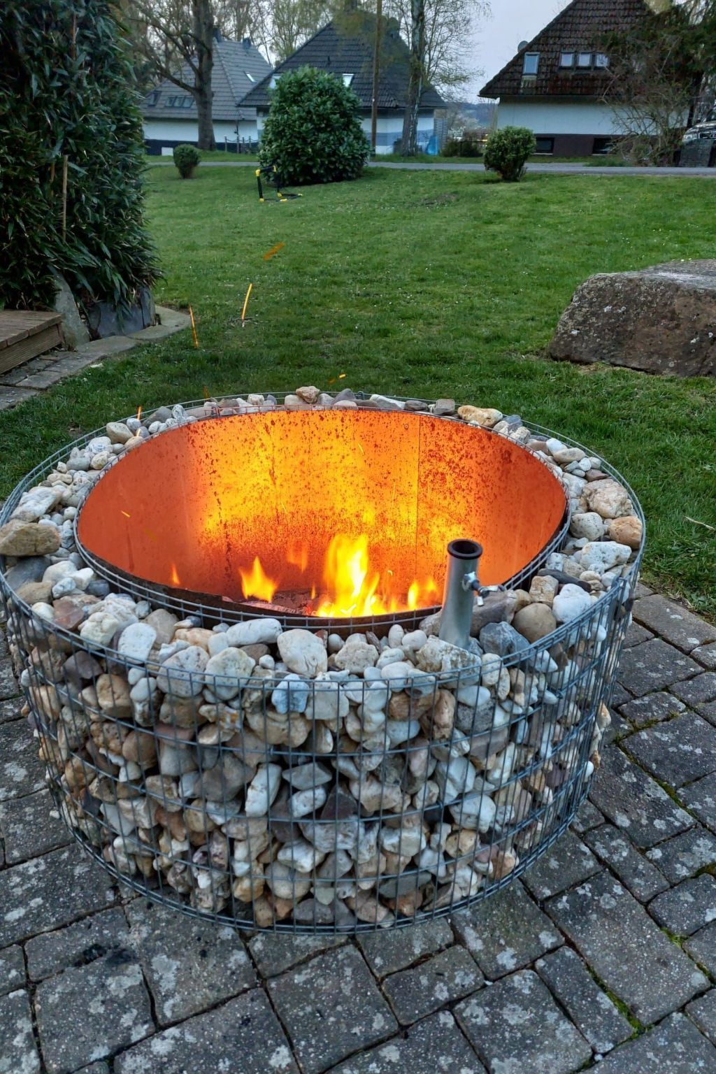A circular outdoor fire pit surrounded by rocks, set on a paved area with a grassy lawn and houses in the background.