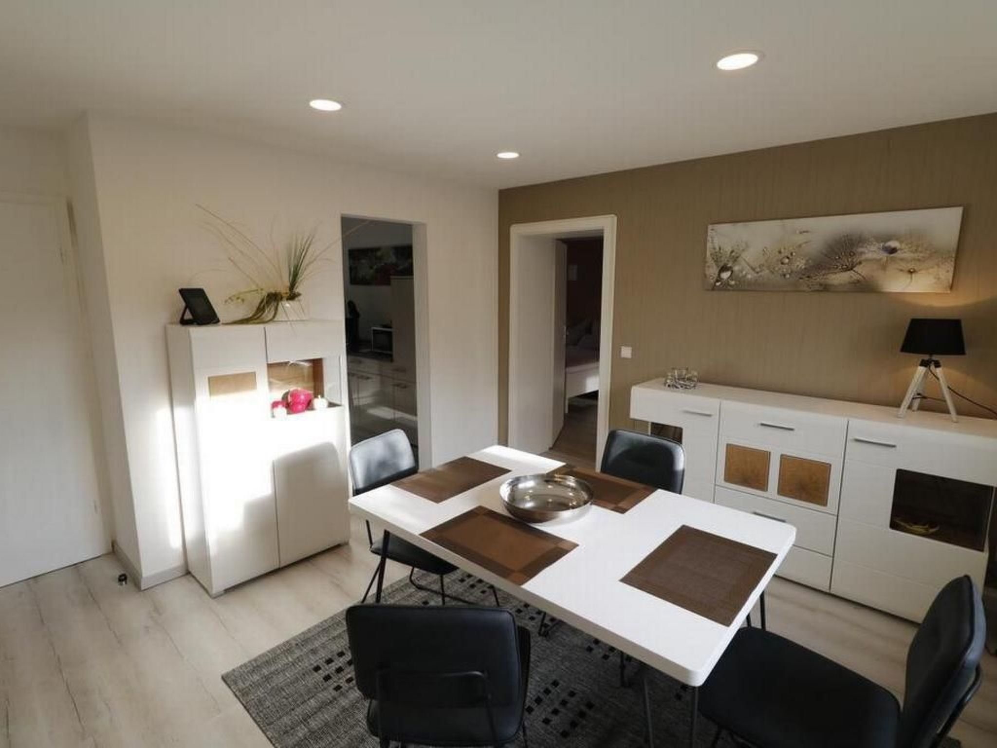 A modern dining area with a white table, black chairs, and a decorative centerpiece. There is a white cabinet with a vase on top and a painting on the wall.