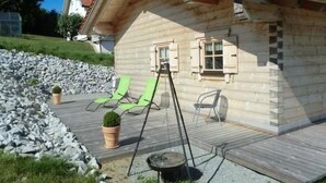 Una cabaña de madera con techo inclinado, una terraza con dos sillones reclinables verdes, una mesita y una planta en maceta.