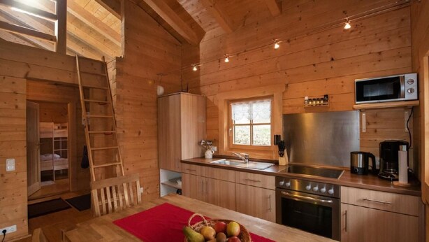 Interior de una cabaña de madera con cocina, comedor y habitación superior con escalera.