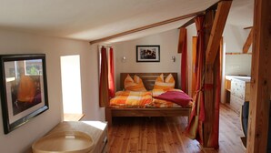 A bedroom with a wooden floor, a bed with orange and white bedding, a wooden headboard, a wooden bench, a framed picture on the wall, and a window with curtains. - "Galerie" by Interhome (Kröslin)