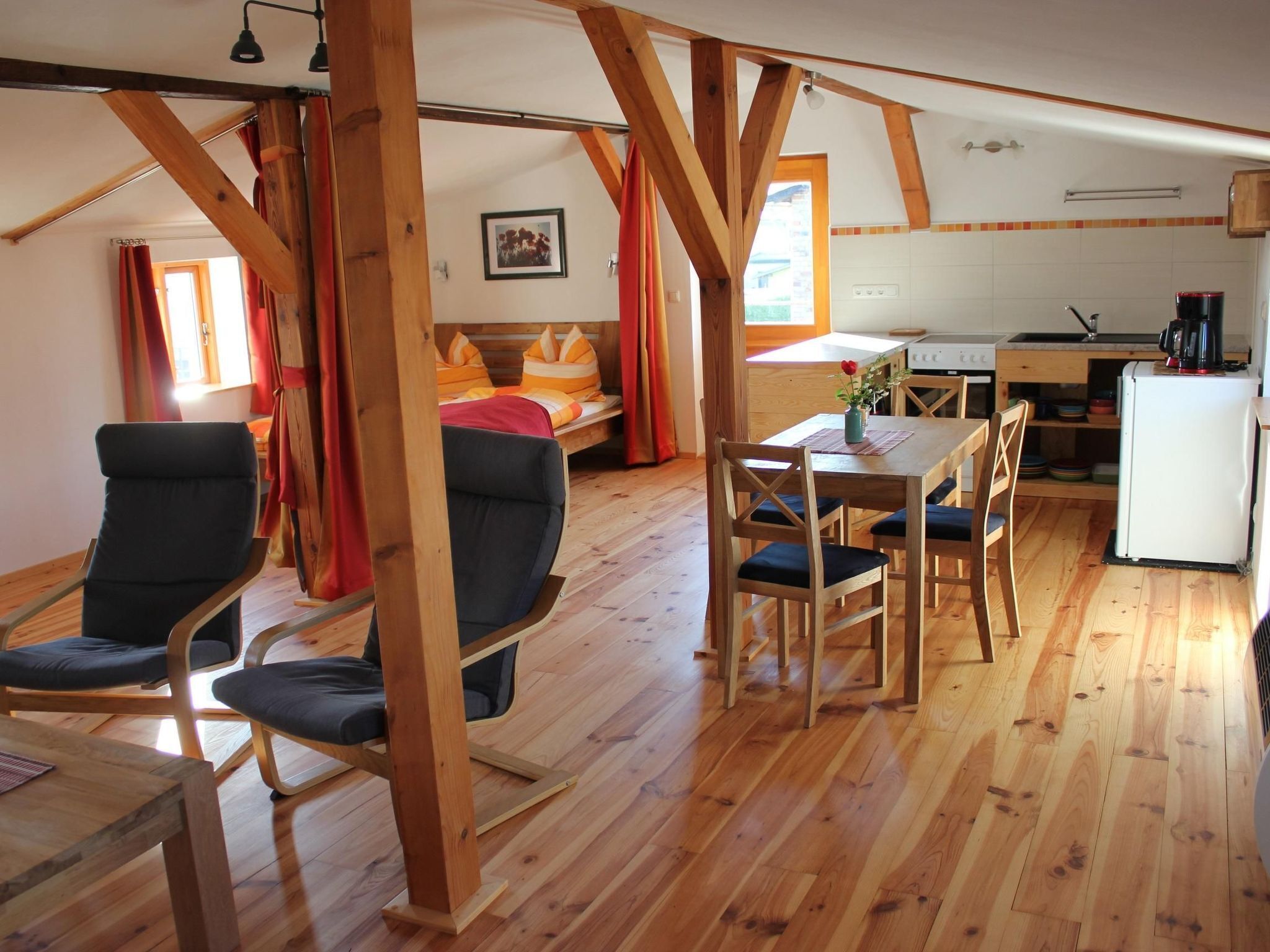 A cozy room with a wooden floor, a bed with orange bedding, a dining table with chairs, and a kitchenette area.