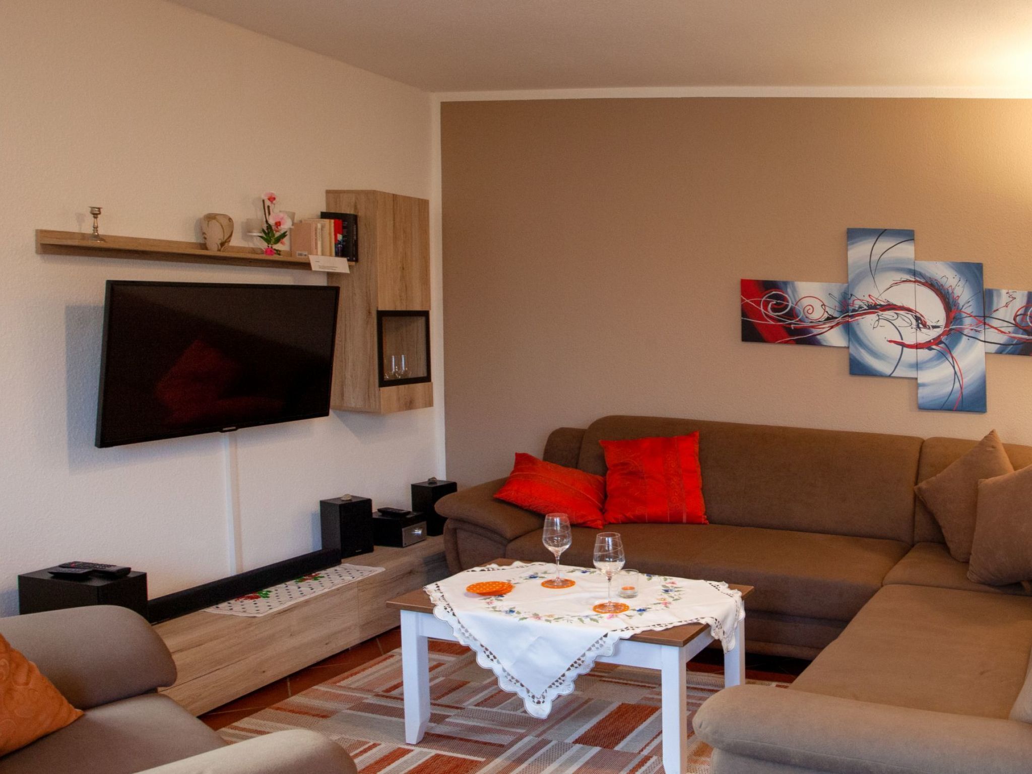 Een woonkamer met een flatscreen televisie, een bruine bank met rode kussens, een salontafel met een witte tafelkleed en abstracte wanddecoratie.